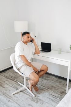 A barefoot man sits in a minimalist home office while talking on a phone and working on a laptop.
