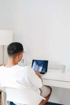 A man sits at a desk using a laptop for online studying, in a bright home interior.