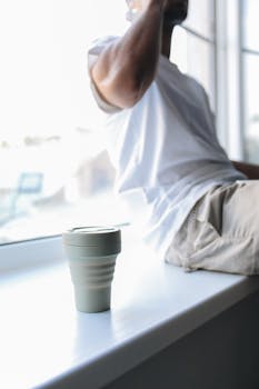 A man sits by a window with a reusable coffee cup, embracing eco-friendly living.