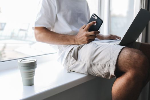 Casual scene of a person using a smartphone and laptop by the window with a cup.