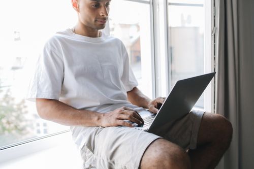 Free A man in a white t-shirt uses a laptop while sitting by a window, focusing on work or study. Stock Photo