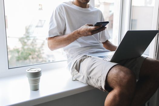 Young man multitasking with smartphone and laptop while sitting by a window.