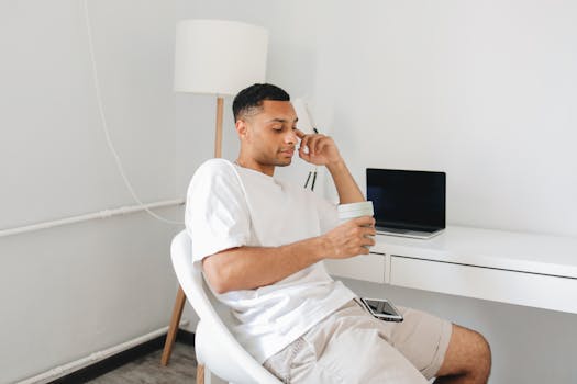 A young man relaxes with coffee in a modern home office, sitting at a white desk with a laptop.