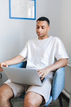 Young man sitting comfortably in an armchair using a laptop at home in a bright room.