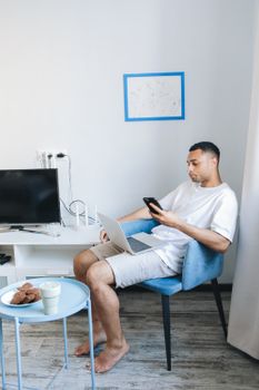 Man sitting at home using smartphone and laptop, relaxing with coffee and cookies.
