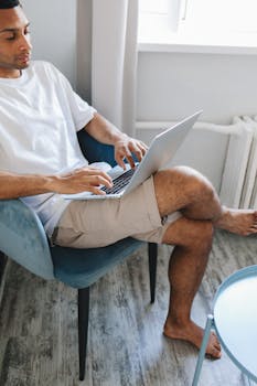 Casual adult man sitting indoors using a laptop with crossed legs in a calm home setting.
