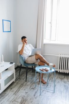 Adult man studying at home with a laptop, sitting on a chair with coffee and croissants on the table.
