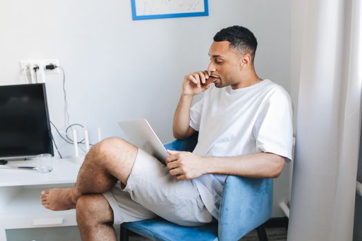 A man in casual clothes working remotely from home, seated in a cozy living room with a tablet.