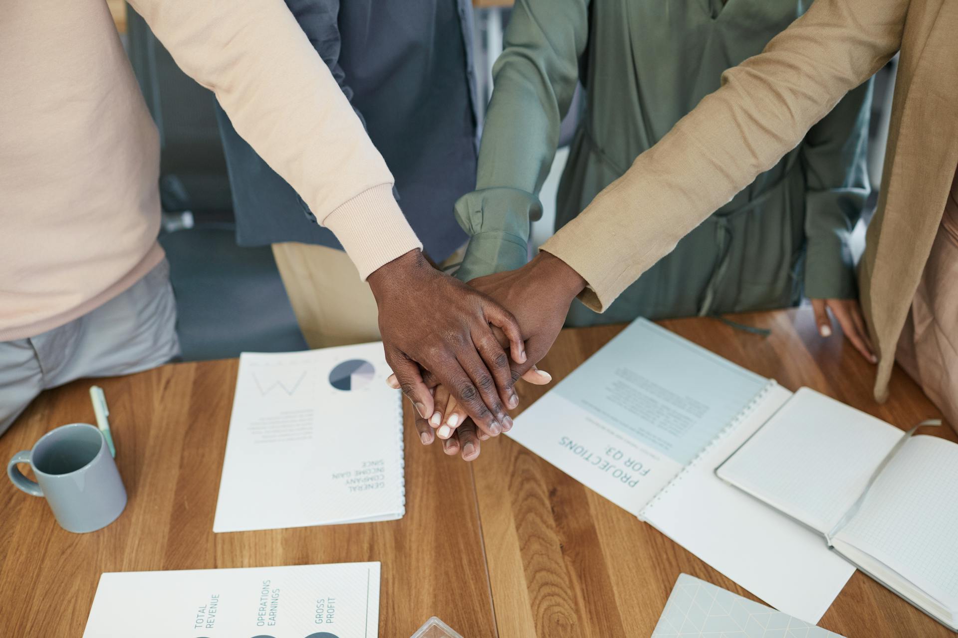 Diverse team stacking hands in unity over a work table, symbolizing cooperation and teamwork.