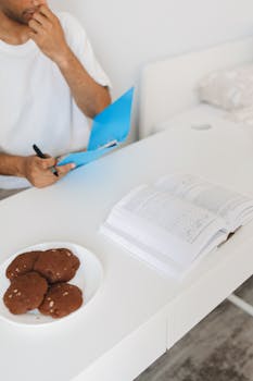 Man seated at white desk with an open book and plate of cookies, studying attentively.