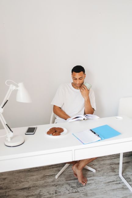 Man seated at home desk reading, surrounded by books, cookies, and a smartphone. Man seated at home desk reading, surrounded by books, cookies, and a smartphone.