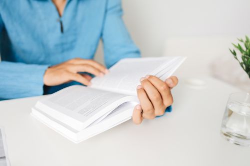 Free Person in a blue shirt studying a book at a white desk indoors with a plant. Stock Photo
