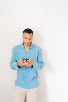 Young man in blue shirt texting on smartphone against white background.