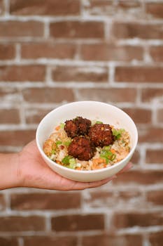 A close-up of falafel balls and salad in a bowl held by a hand in front of a brick wall.