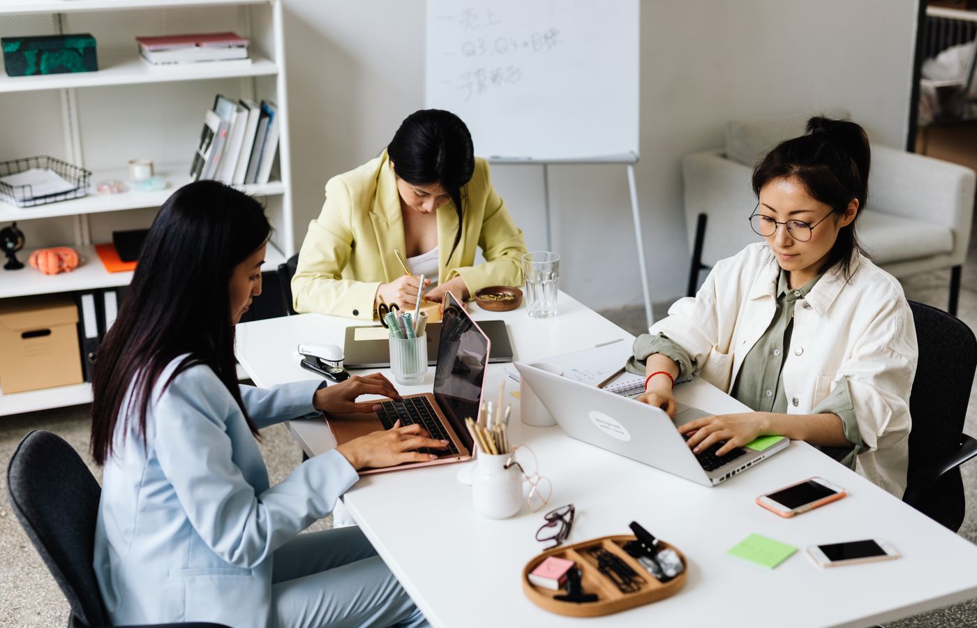 Three Asian women focused on laptops and notes, collaborating in a modern office setting.