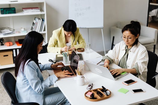 Three Asian women focused on laptops and notes, collaborating in a modern office setting.