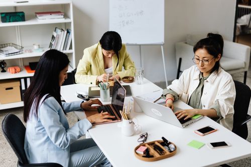 Free Three Asian women focused on laptops and notes, collaborating in a modern office setting. Stock Photo