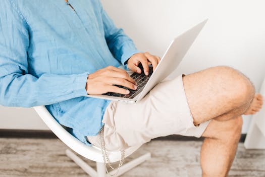 A person in casual shorts and top typing on a laptop while sitting indoors.