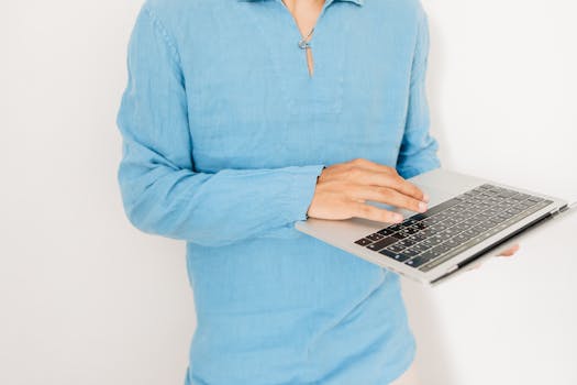 Close-up of a person using a laptop, wearing a casual blue shirt against a plain background.