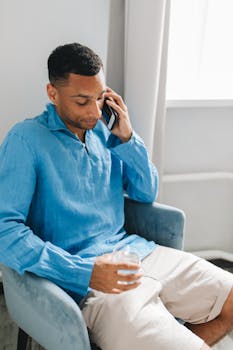 Young man in a blue shirt talking on his phone while seated indoors.