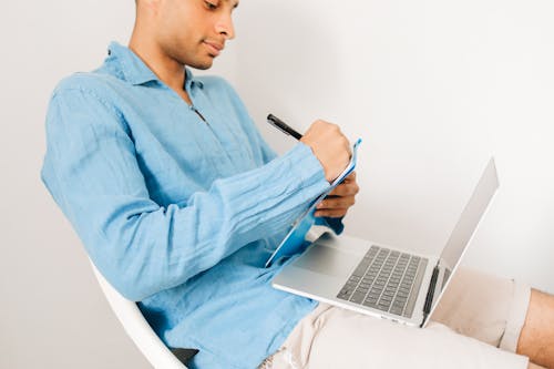 Close-Up Shot of Man in Blue Long Sleeves Writing with a Laptop on His Lap
