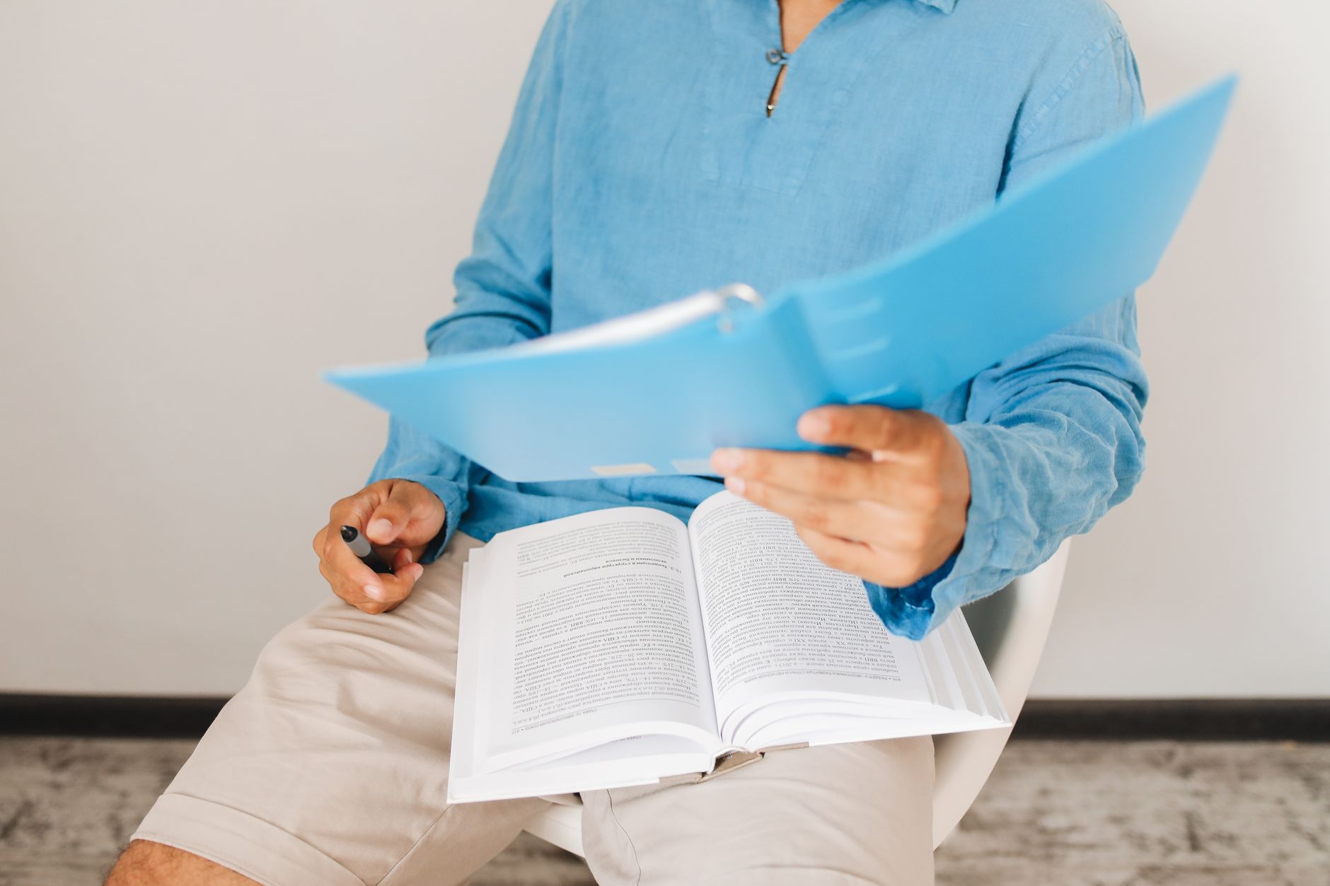 A man in casual attire reads and takes notes from a book indoors, adding insights.