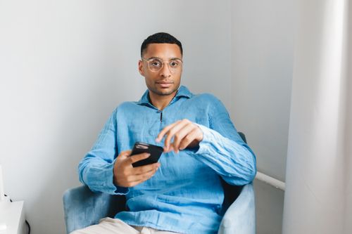Free Casual portrait of a man in glasses sitting indoors wearing blue and holding a smartphone. Stock Photo