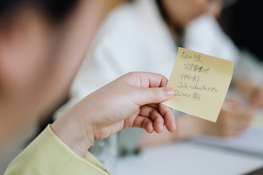 A close-up of a hand holding a yellow sticky note with handwritten text during a meeting.