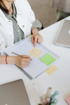Woman writing on a spiral notebook, planning with sticky notes at a desk.