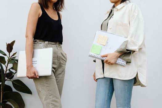 Two women standing and holding a laptop and documents in an office setting.
