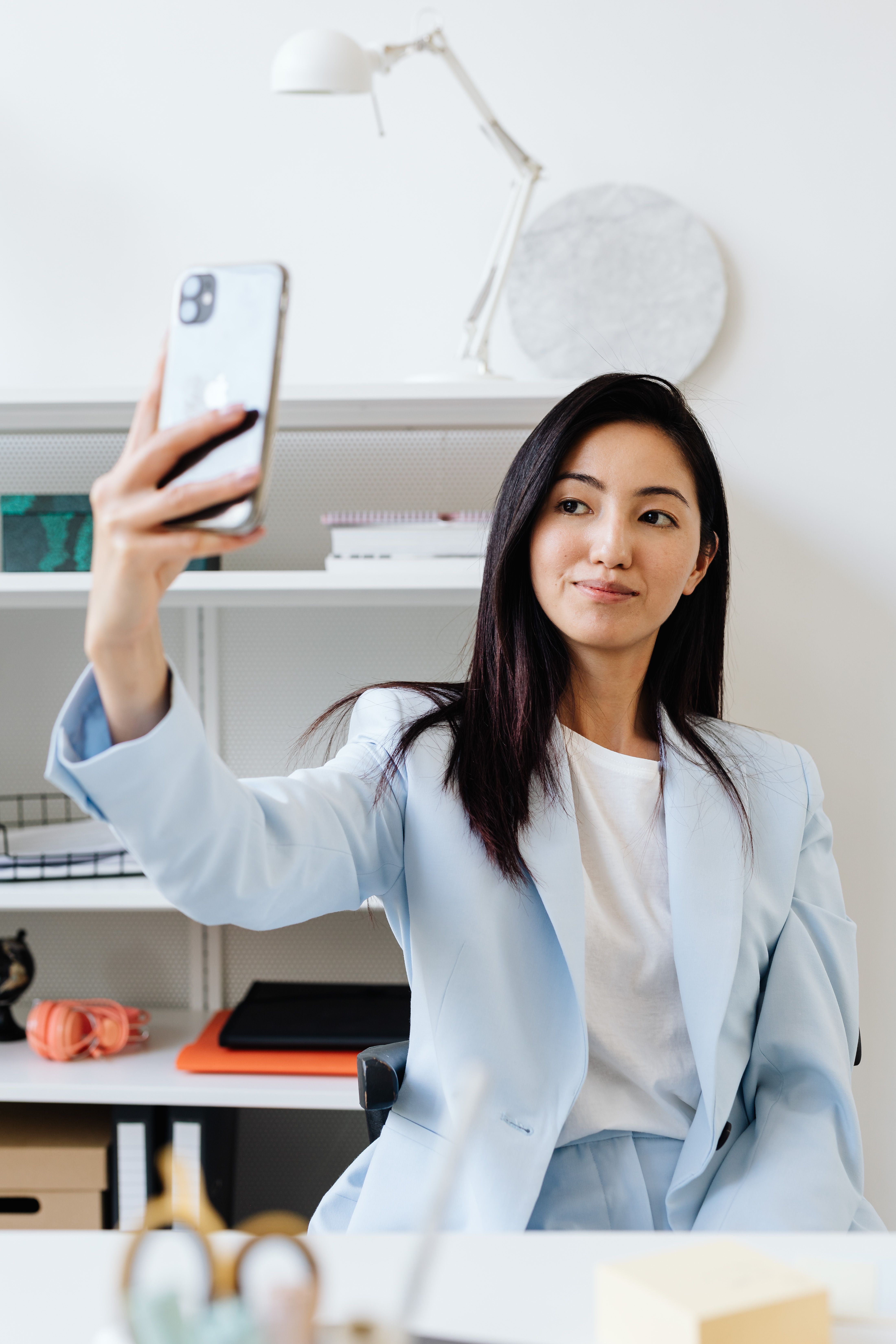 Asian woman in blue suit takes a selfie in a modern office setting.