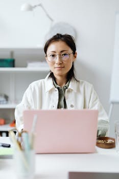 Young woman in glasses working at a desk with a laptop in a modern office setting.