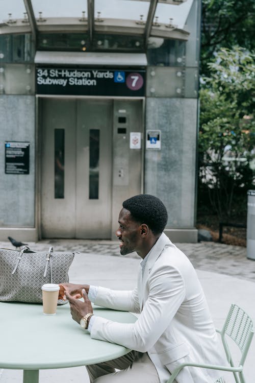 Elegant Man Sitting at a Table in Front of an elevator to a Subway Station and Drinking Coffee 