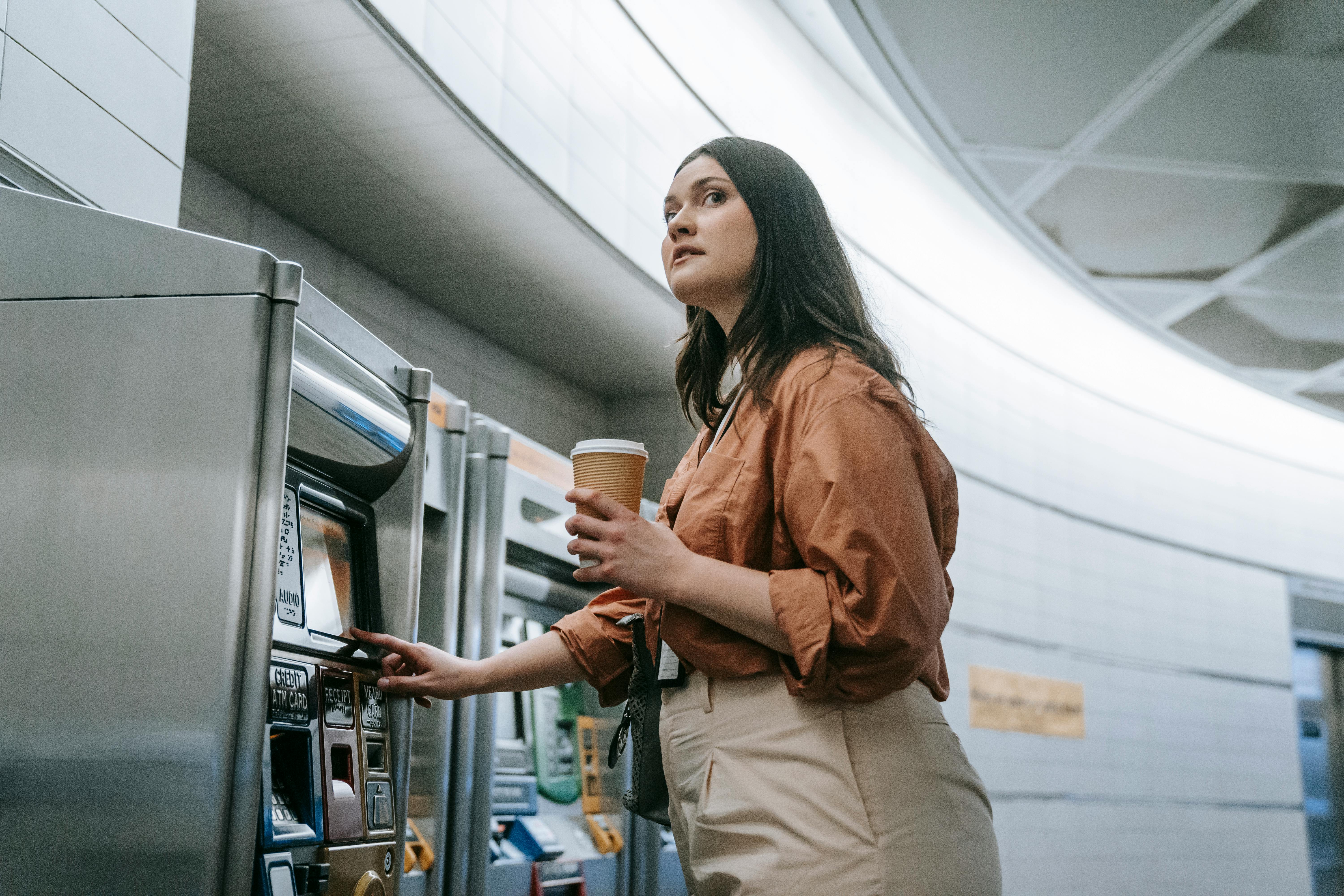 Woman in subway station using a ticket machine while holding a coffee cup.
