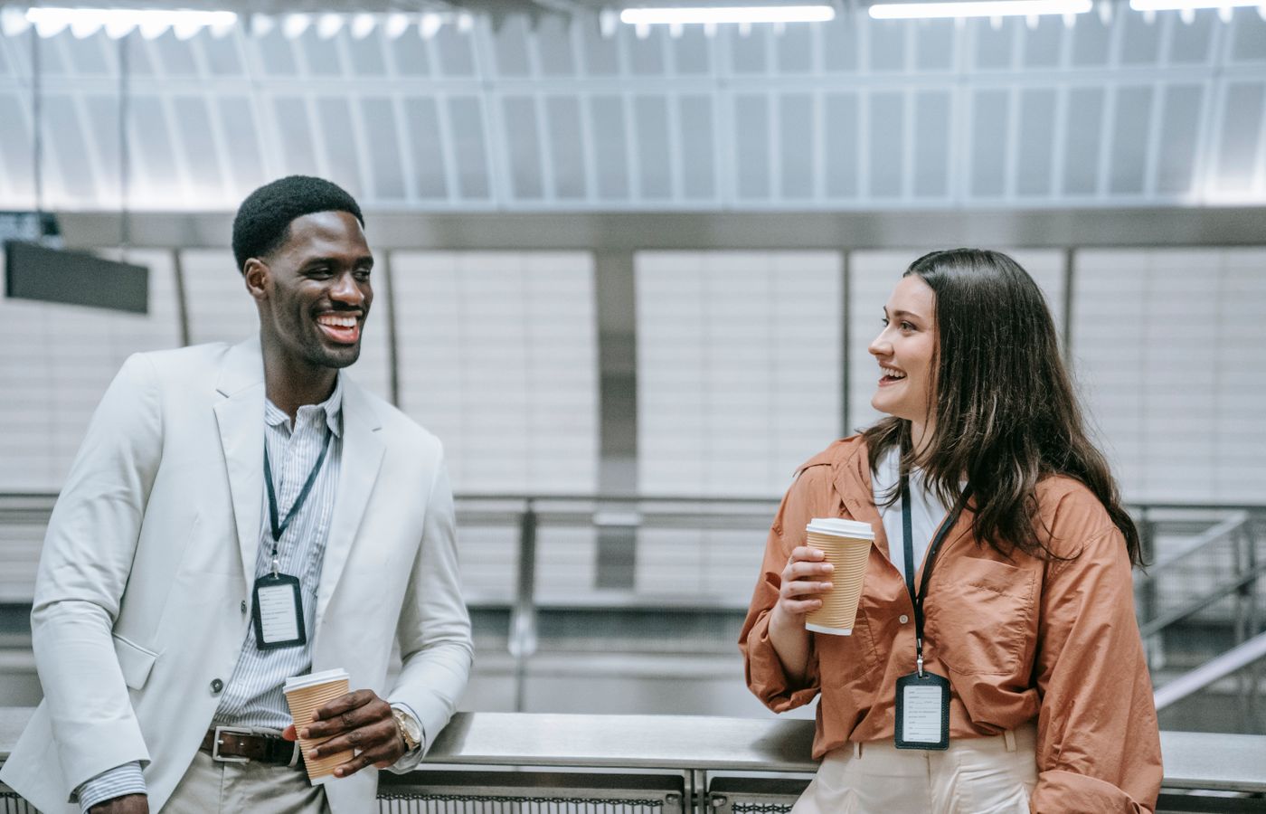Two smiling coworkers with ID badges enjoy a coffee break indoors.
