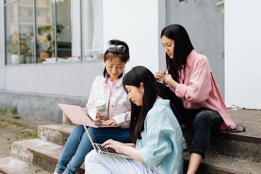 Three young women sitting on outdoor steps, collaborating on laptops.