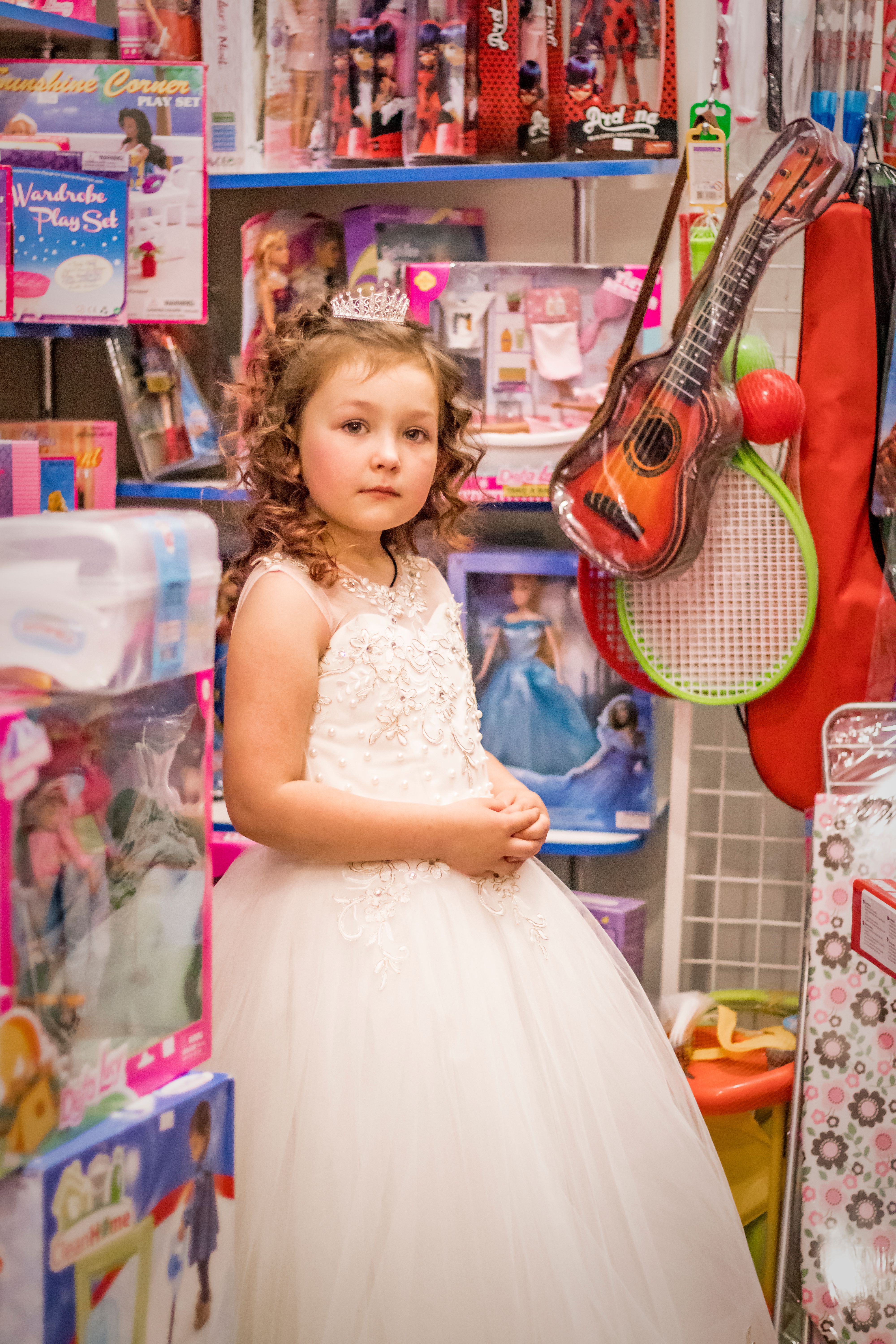 A young girl in a white gown and tiara stands in a colorful toy store setting.