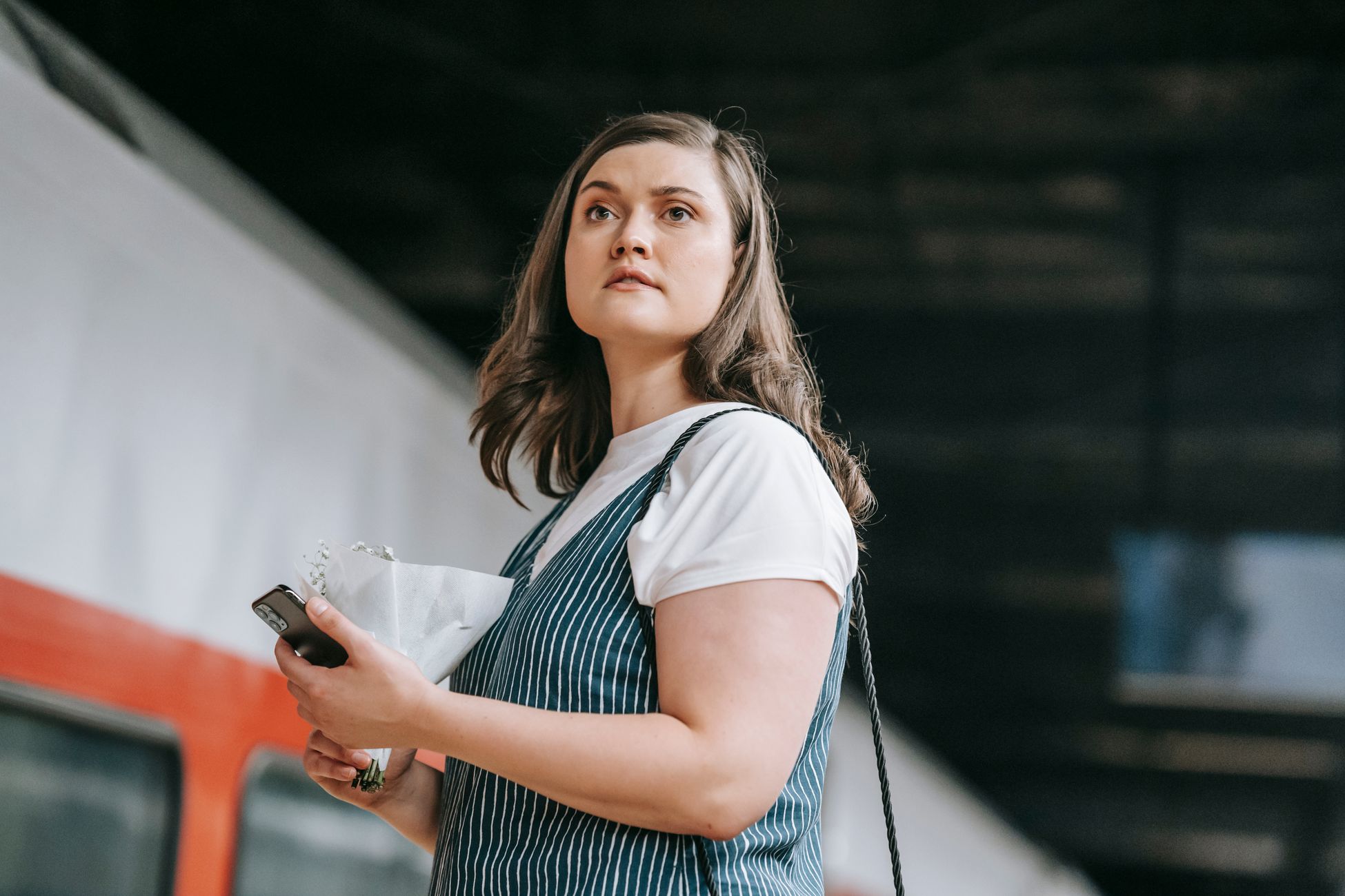 Young woman using phone at a train station, waiting for departure under dim lighting.
