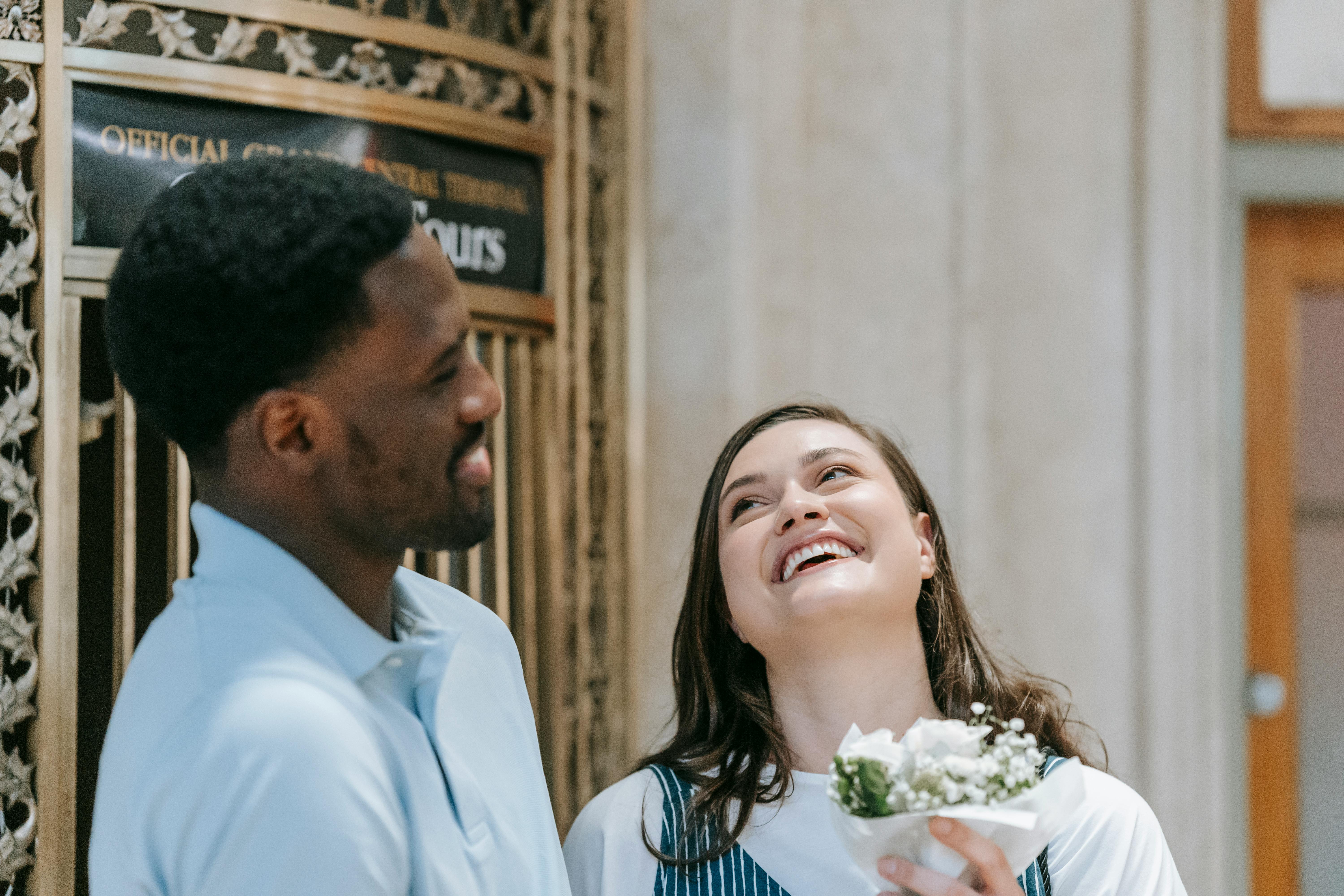 Joyful couple smiling while holding a bouquet indoors, capturing a happy moment.