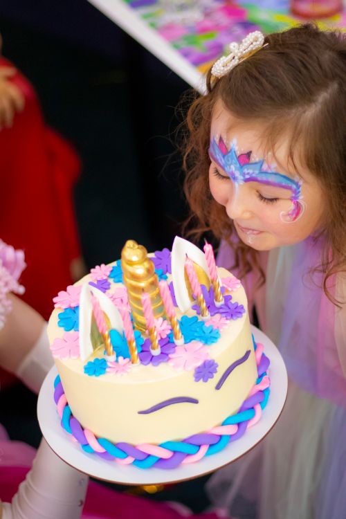 Free A young girl with face paint leans over a colorful unicorn-themed birthday cake, ready to blow out the candles. Stock Photo