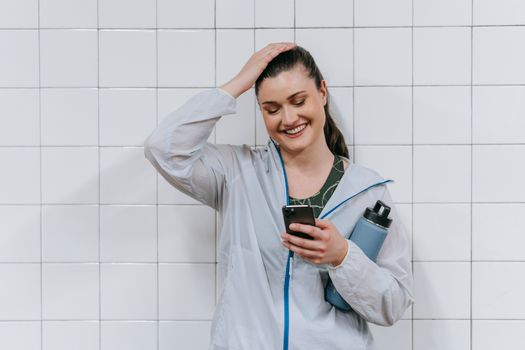 Caucasian woman in sporty attire smiling at smartphone with tumbler indoors.