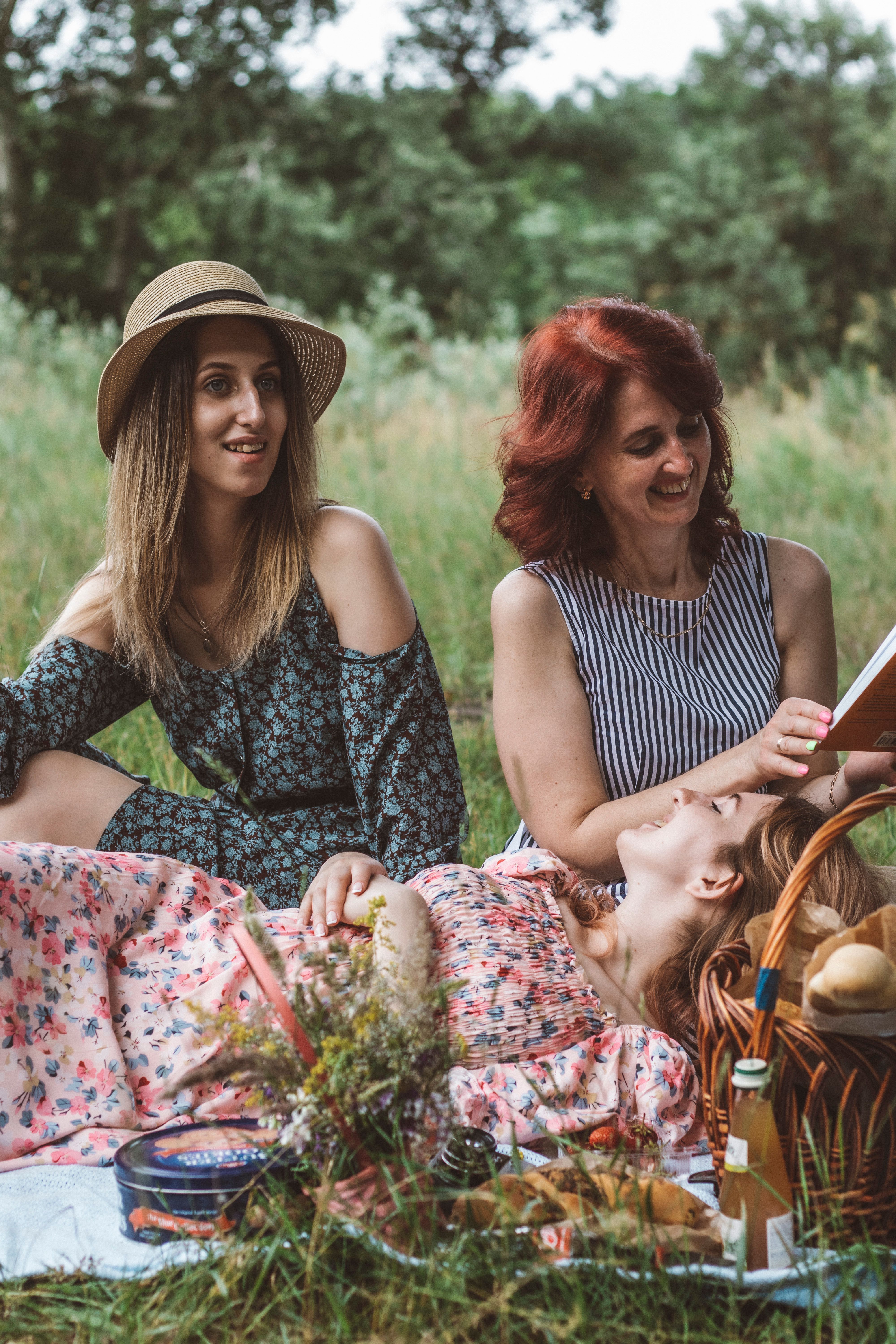 Three women enjoying a summer picnic with food and laughter in a tranquil outdoor setting.