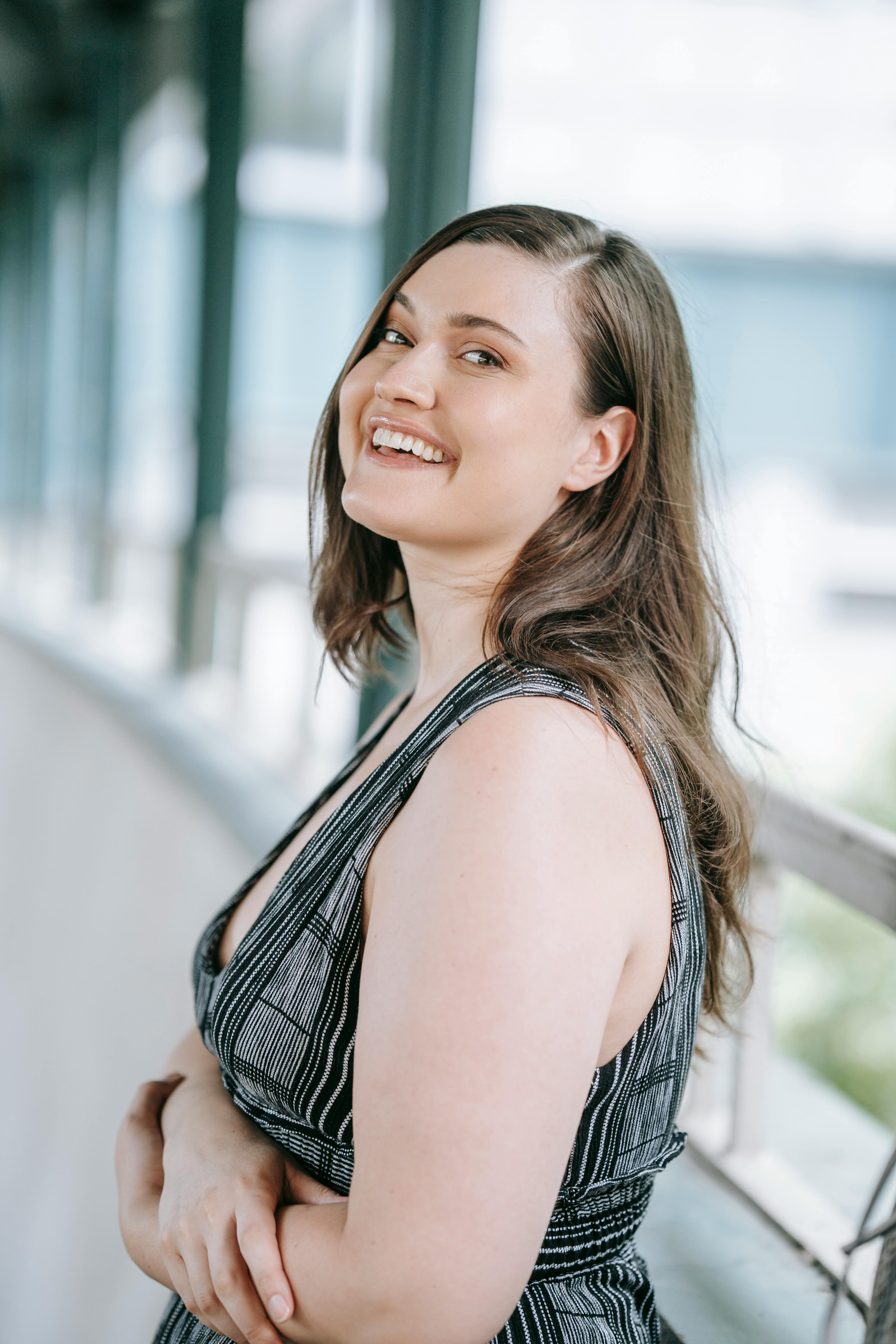 A happy woman with long hair smiling in a striped dress, posing on a walkway outside.