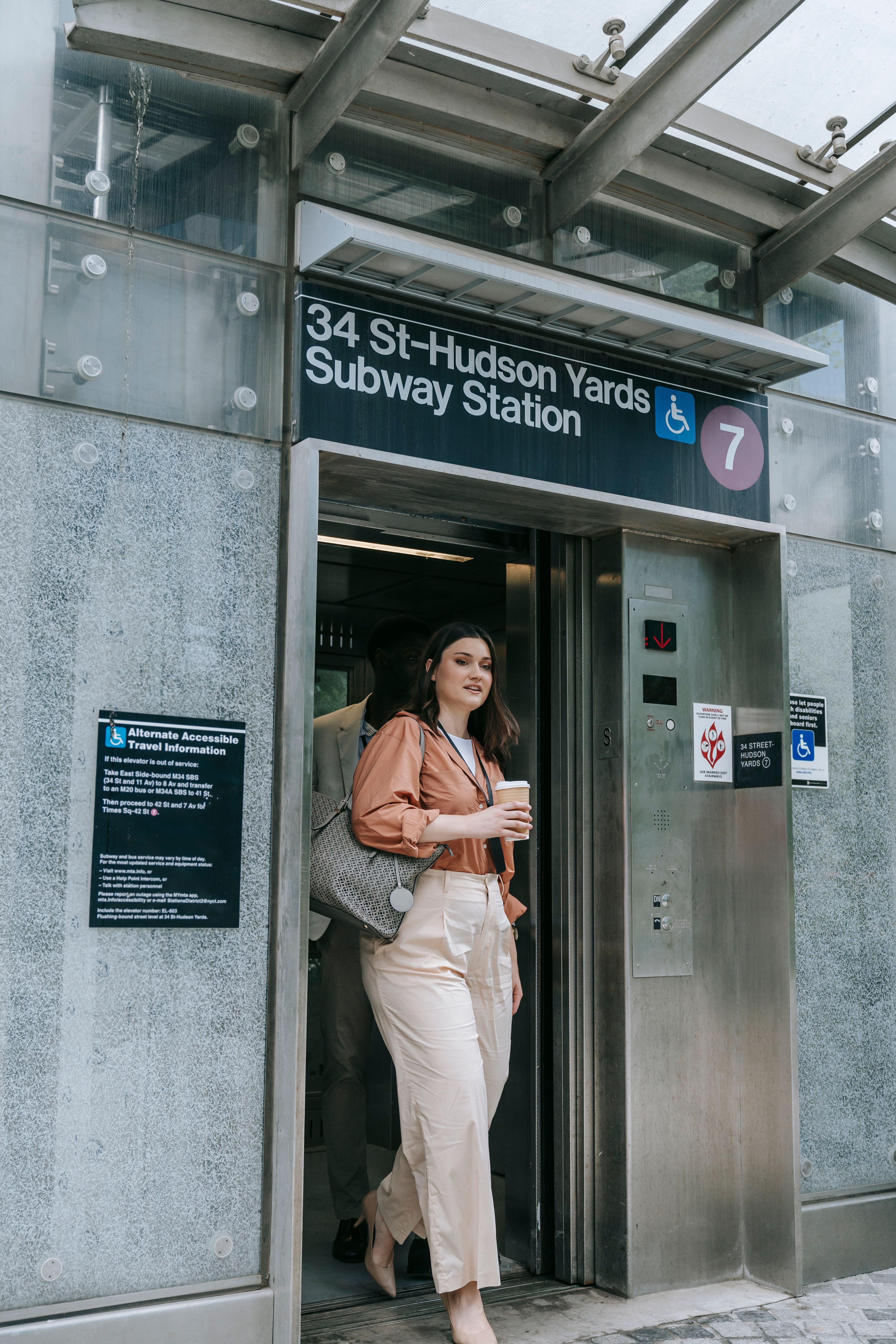 Stylish woman holding coffee exits elevator at 34th St-Hudson Yards Subway Station.