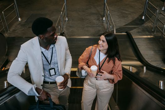 Business professionals with coffee cups on escalator, engaging in light conversation.