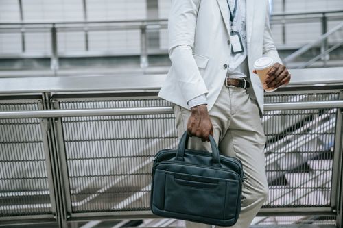 Free A stylish businessman in a white suit holds a leather bag and a coffee, leaning against handrails indoors. Stock Photo