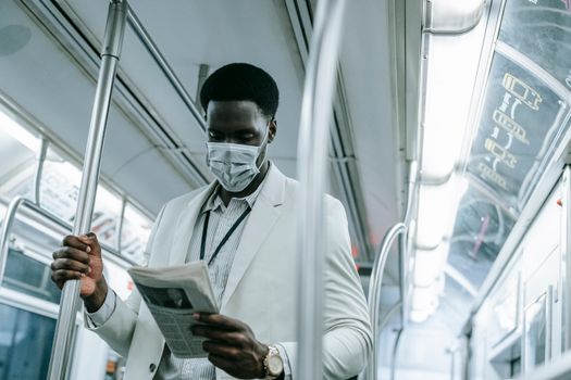 A man in a mask reads a newspaper while standing on a subway train.