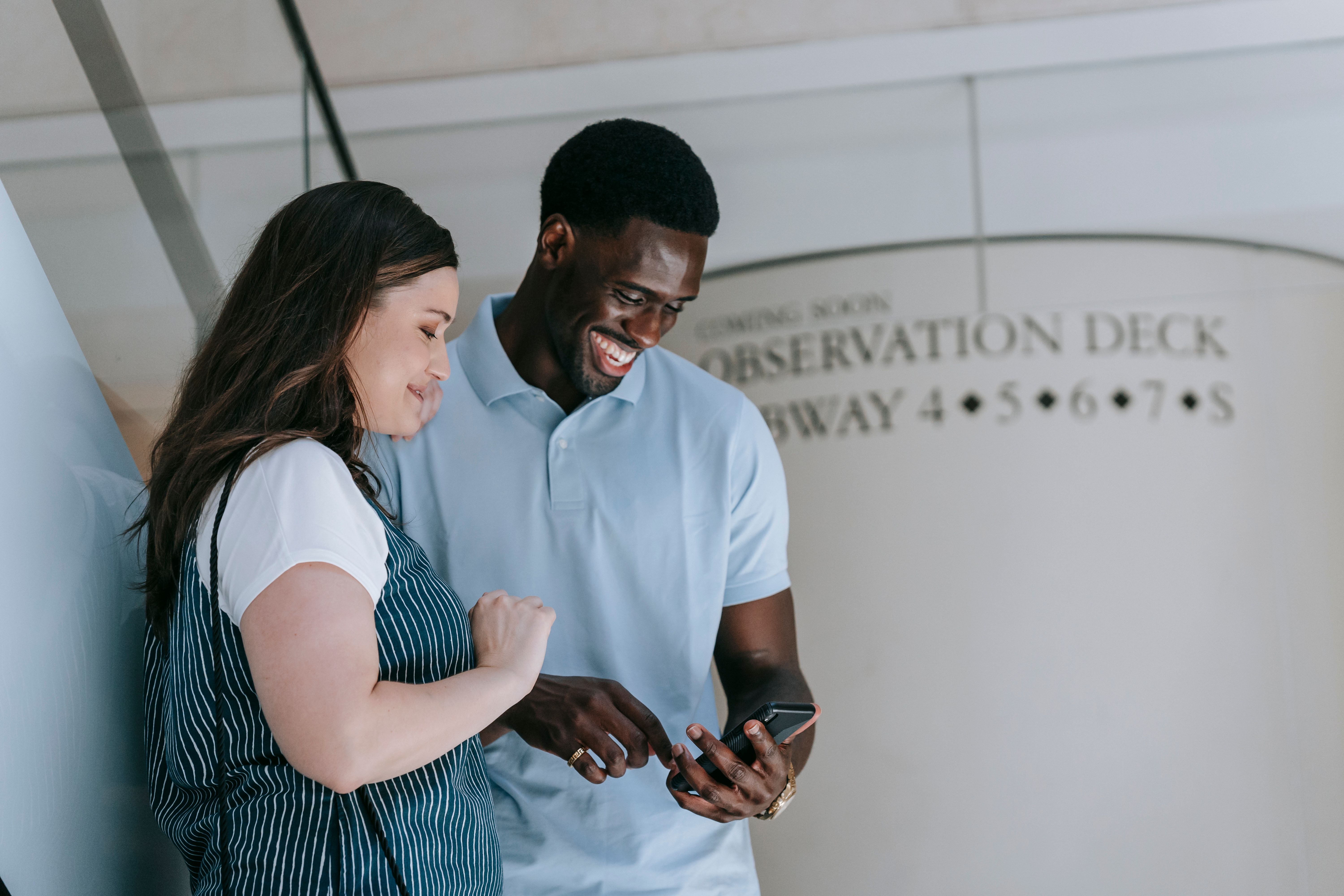 Two friends laughing and sharing a moment while looking at a smartphone indoors near an observation deck.