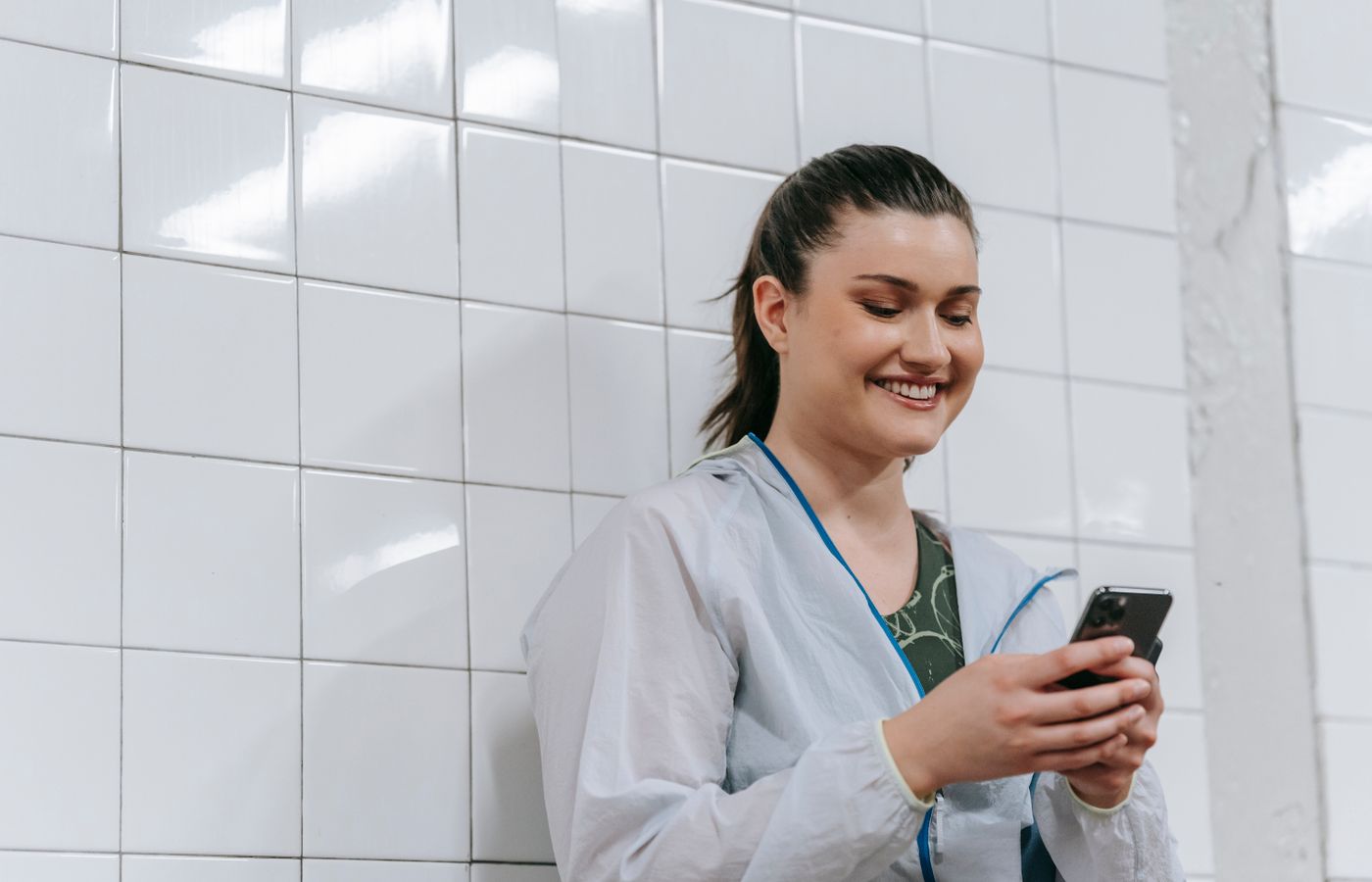 A woman happily texts on her smartphone against a white tiled wall indoors, wearing a casual jacket.