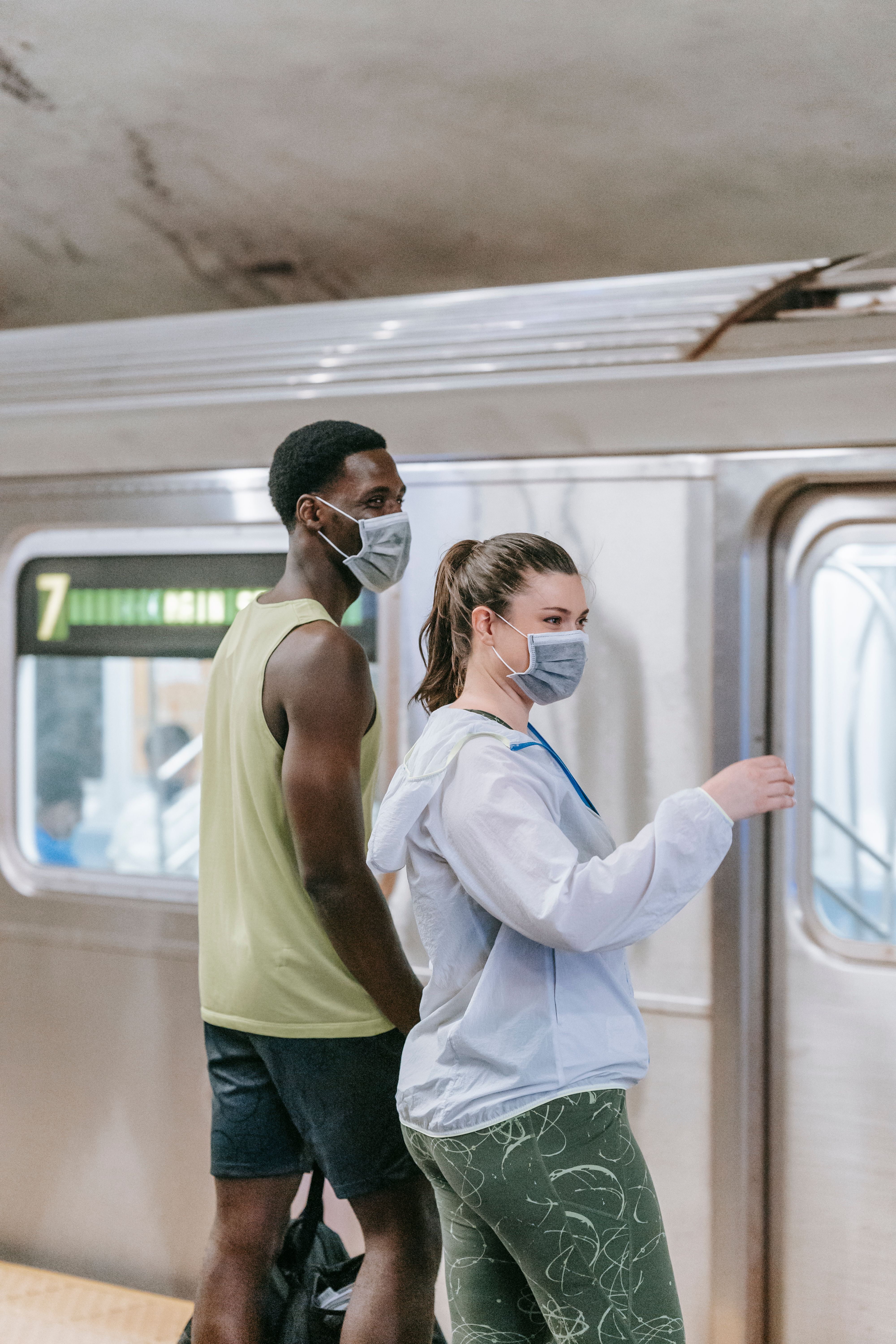 Two adults wearing masks stand at a train station. Captured in a vertical composition.